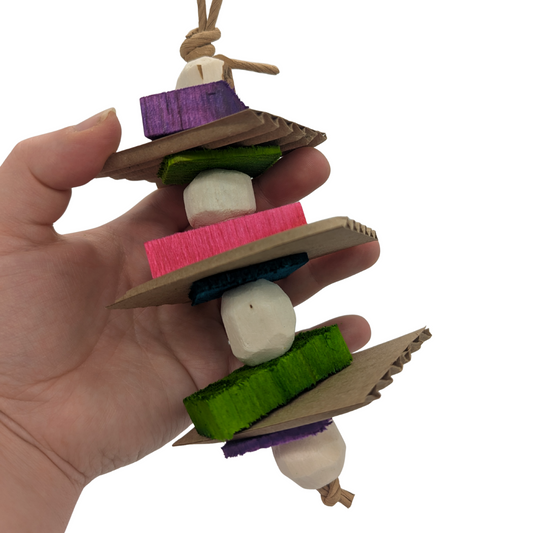 A hand holding bird toy with alternating white sola balls, cardboard slices, colorful balsa slats, and 1/8" pine slices on a white background.