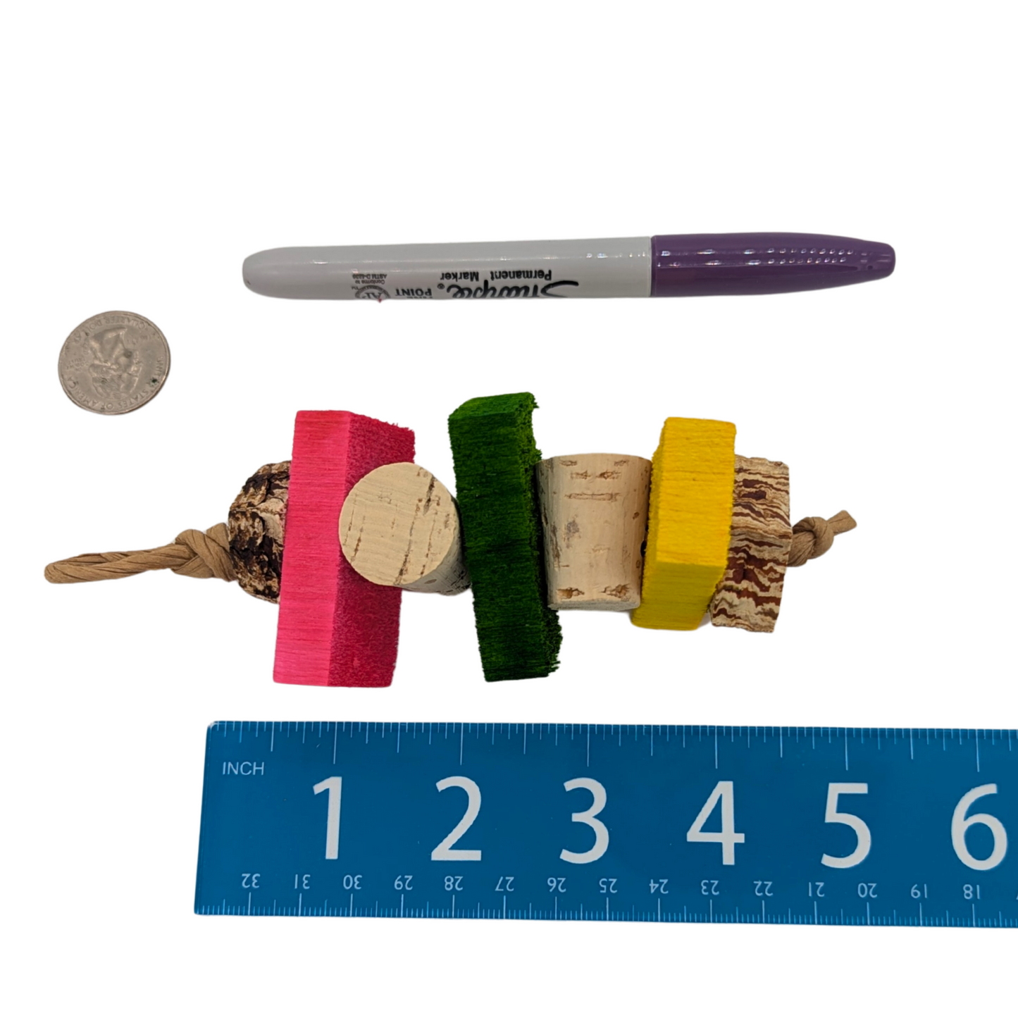 Colorful bird toy with natural elements next to a pen and ruler for scale on a white background