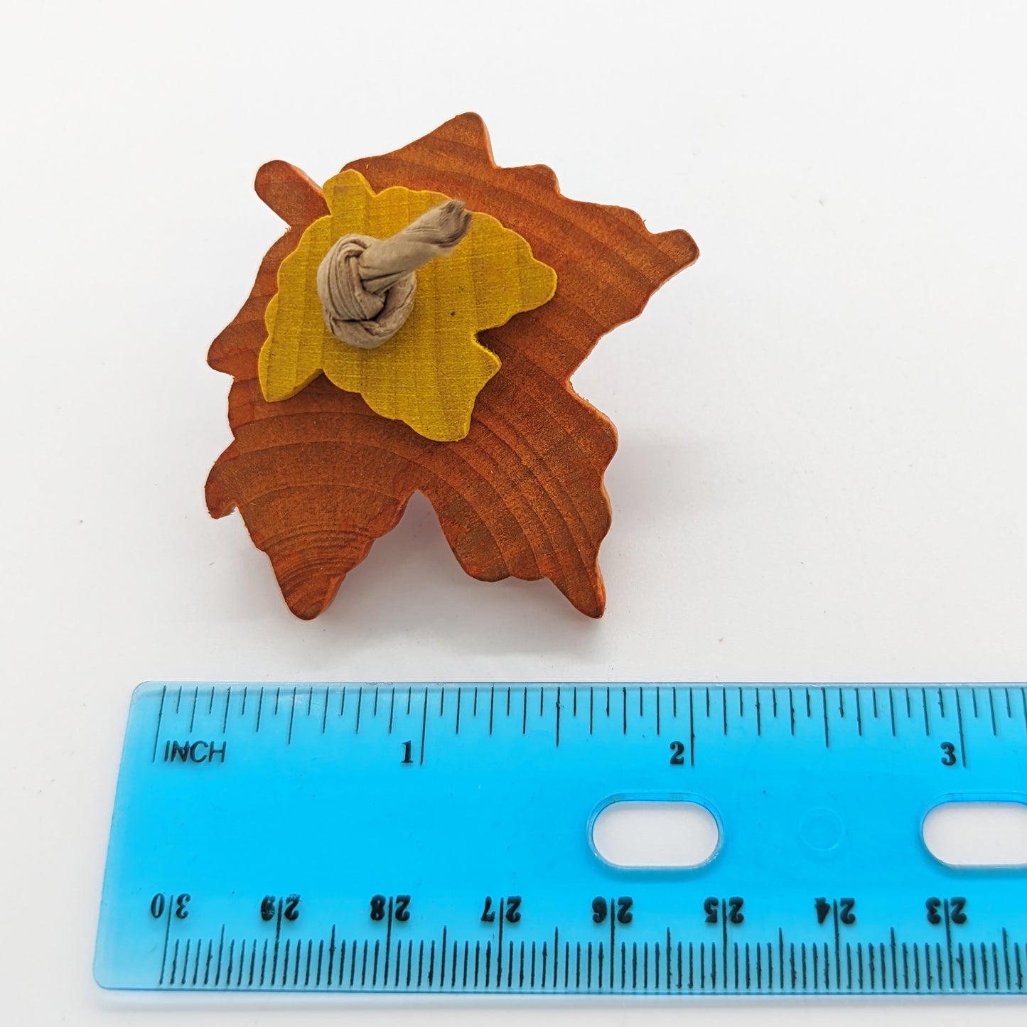 A orange wooden maple leaf bird toy, with a smaller yellow maple leaf tied on it with paper rope. Shown with a ruler to show size (about two inches wide).