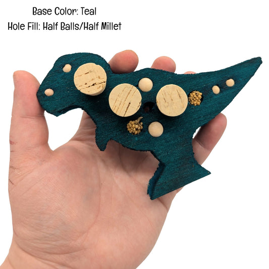 Balsasaurus Rex by Lil Monsters Bird Toys. A piece of balsa cut into the shape of a t-rex dinosaur. Embedded with cork stoppers, millet, and wooden beads. Shown in a teal color, held in hand for scale. 