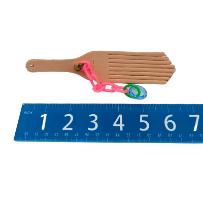 A bird toy with a leather base and plastic additions shown next to a ruler on a white background. 