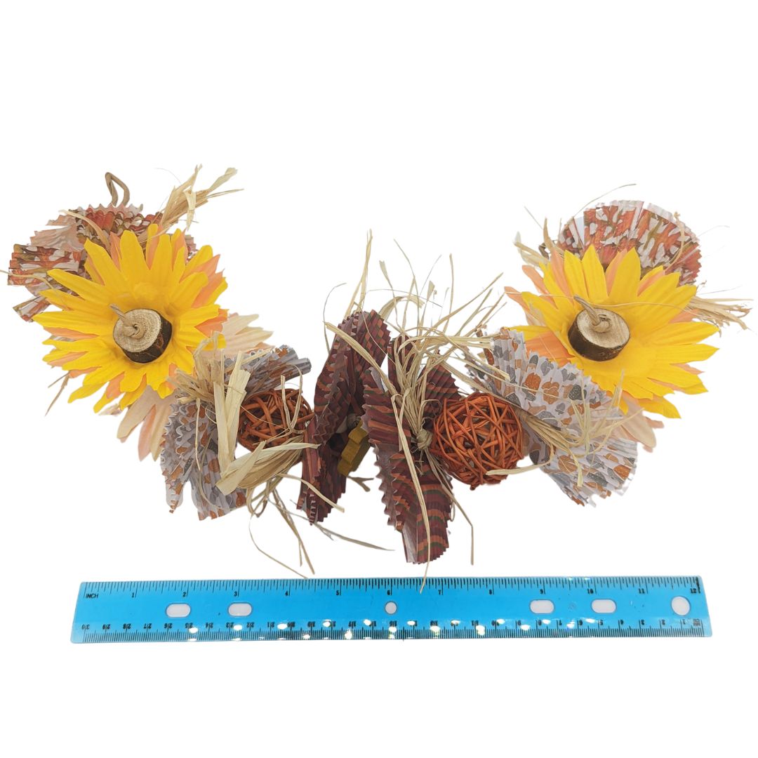 A fall themed bird toy that hangs from both ends like a garland. Has raffia, balsa blocks, and cupcake liners cut and layered to resemble sunflowers. Shown with ruler.