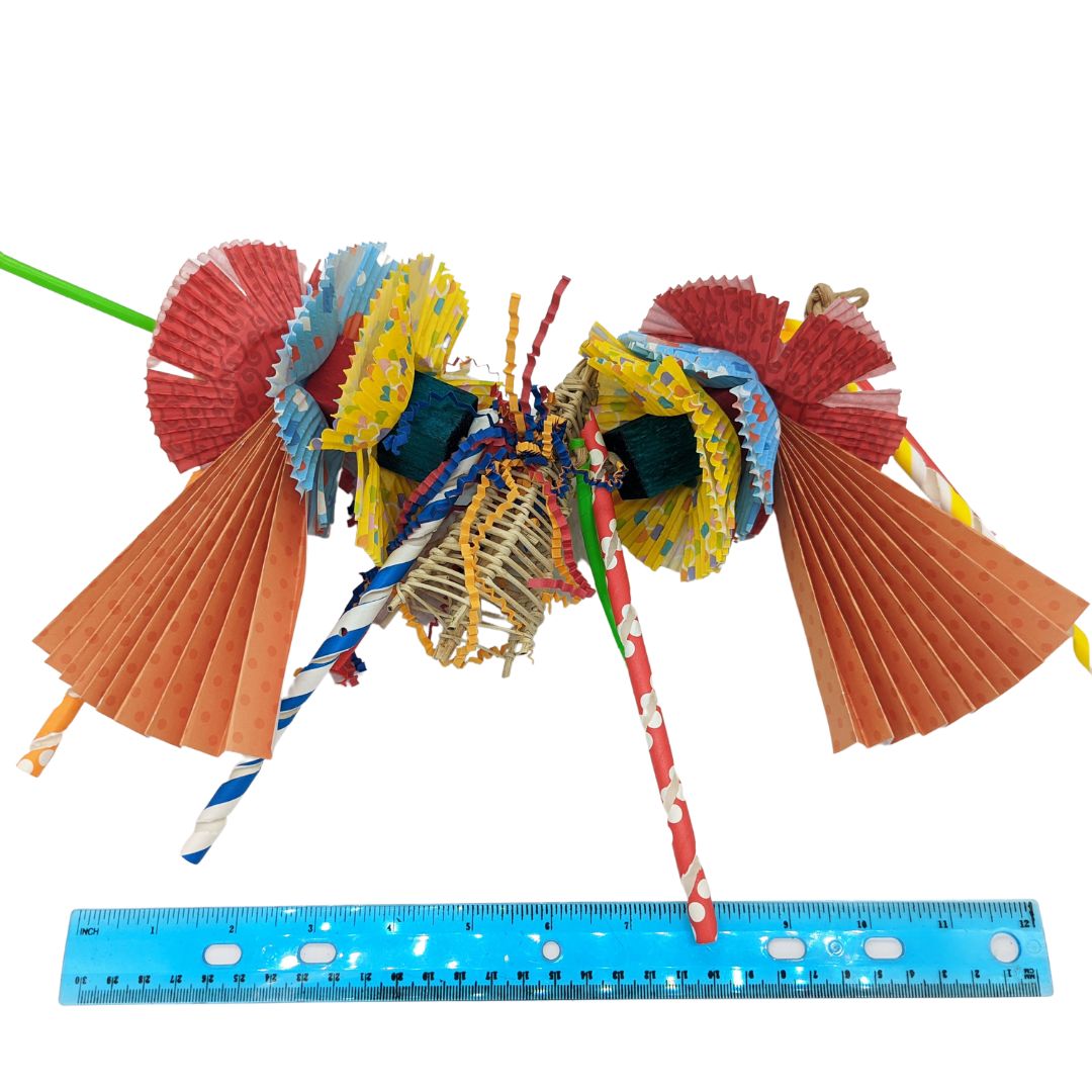 A garland style toy for parrots with cupcake liners, paper straws, and shredded paper, in pink, purple, and yellow colors with various patterns. Includes balsa blocks, a vine party hat, and strung on paper rope. Multicolor coloration. Next to 12 inch ruler